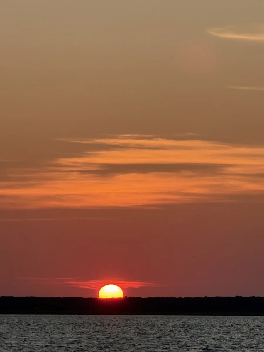 The sun on the horizon beyond the far shore, seen across open water under a pink sky